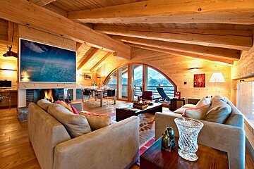 A warm, rustic wooden chalet living room with exposed beams, a cozy fireplace, and large arched windows offering a mountain view. Sofas and dining area visible.