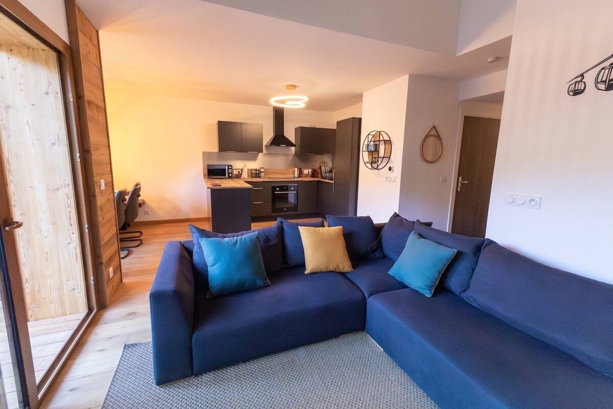A modern open-plan living room with a blue sectional sofa, a sleek gray kitchen, wooden floors, and ski lift wall decor. A balcony door is visible.