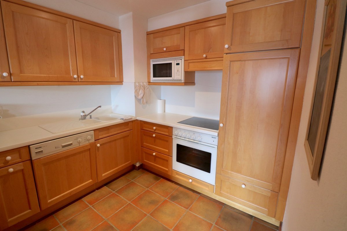 A modern kitchen with light wood cabinets, white countertops, built-in appliances (microwave, oven, dishwasher), and a tiled floor.