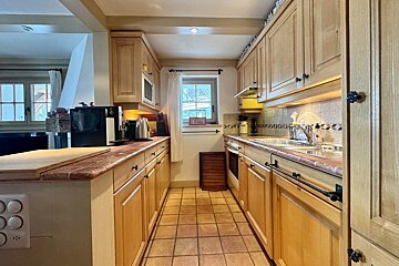A warm, rustic galley kitchen with light wooden cabinets, tiled counters, a terracotta-tiled floor, and a window in the center.