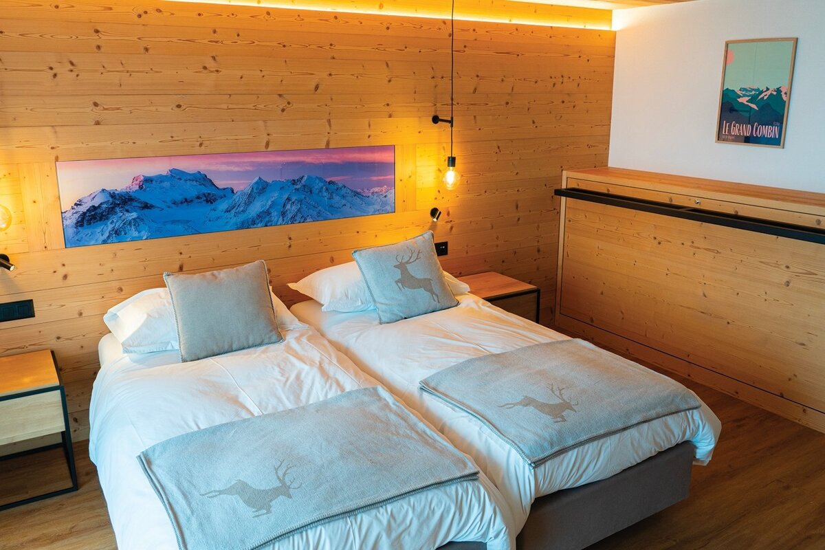 A cozy, modern bedroom features wooden walls, two beds with deer-embroidered throws, a panoramic mountain mural, and warm accent lighting.