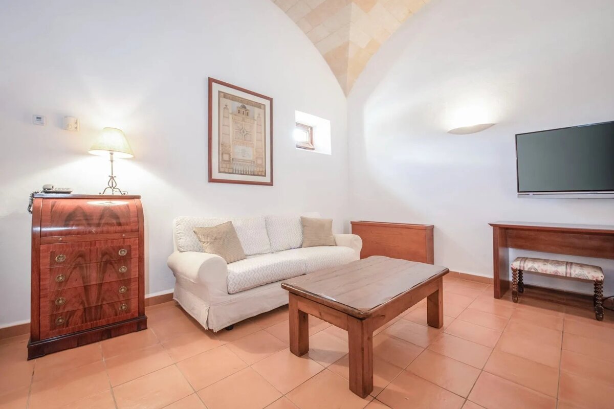 A white-walled living room with a vaulted ceiling, white sofa, wooden coffee table, desk, wall-mounted TV, and terracotta tiled floor.