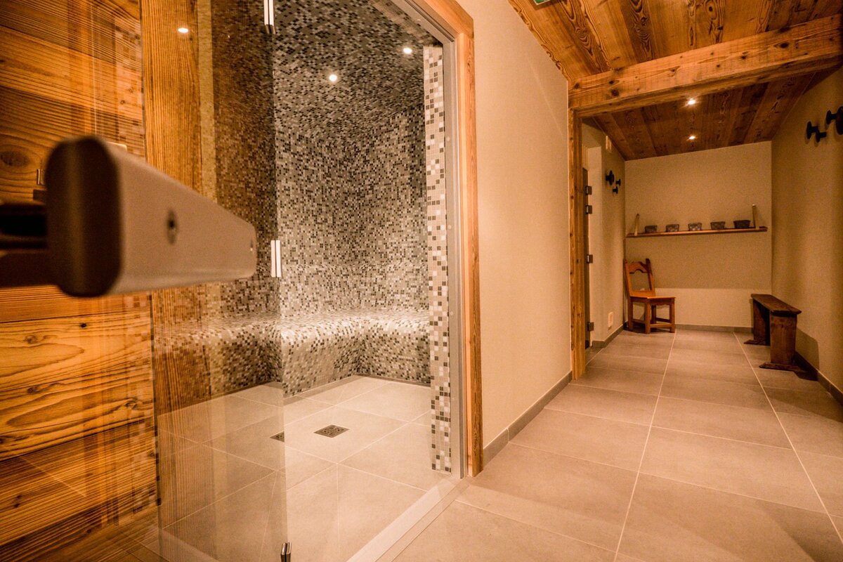 An inviting steam room with mosaic tiles and a glass entrance, adjacent to a warm, rustic hallway featuring a wooden ceiling, chair, and bench.