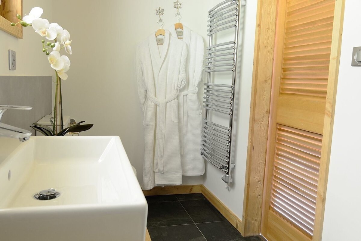 A modern bathroom features a white sink with orchids, two white bathrobes hanging, a chrome towel warmer, a wooden louvered door, and dark tiled floor.