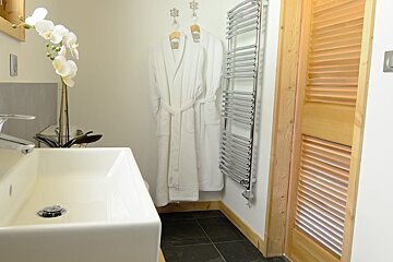 A modern bathroom features a white sink with orchids, two white bathrobes hanging, a chrome towel warmer, a wooden louvered door, and dark tiled floor.