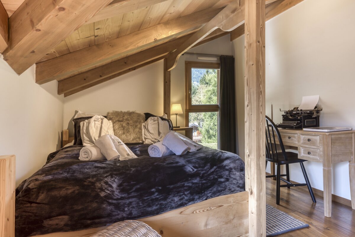 A cozy attic bedroom with exposed wooden beams, a large bed, a desk featuring a vintage typewriter, and a window overlooking a lush forest.
