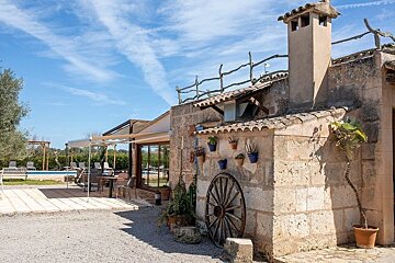 A charming rustic stone house with a wagon wheel, potted plants, and a patio with outdoor seating, overlooking a pool under a bright blue sky.