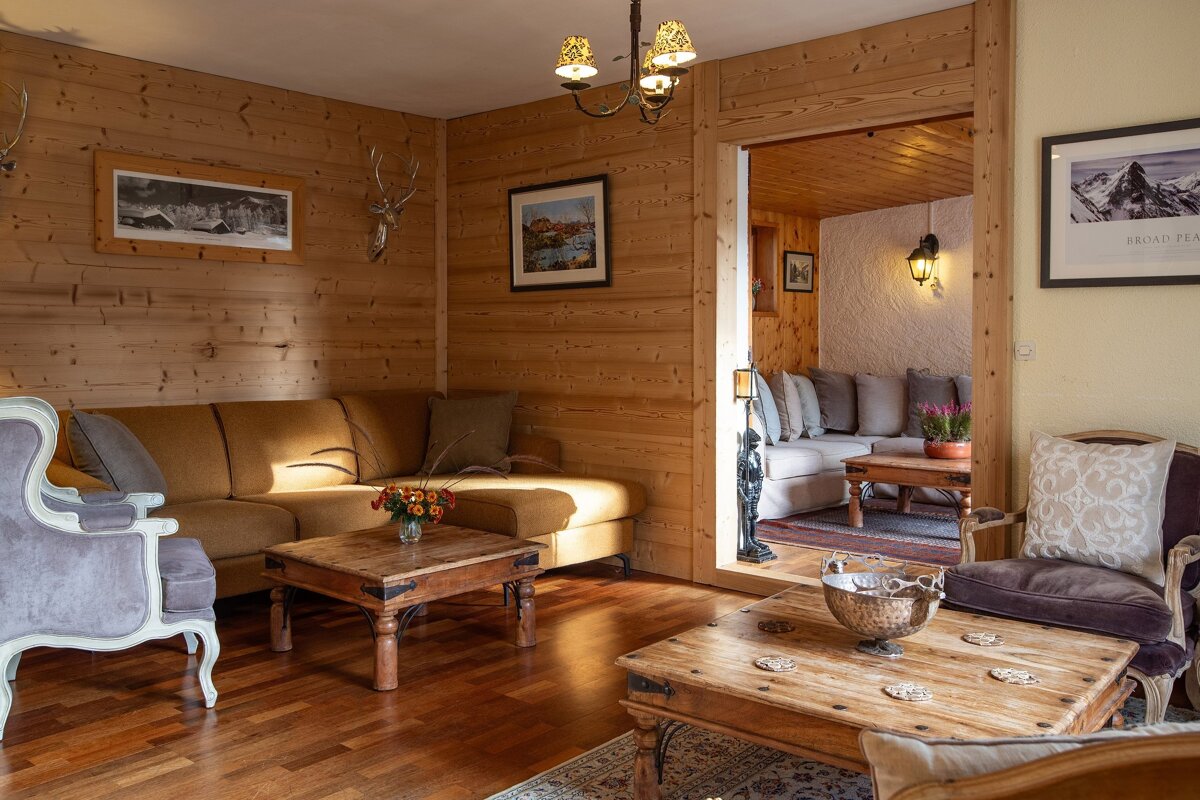 A warm, wood-paneled living room with multiple seating areas, rustic wooden coffee tables, deer head decor, and inviting lighting, leading to another cozy room.