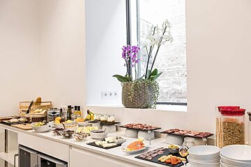 A full breakfast buffet on a white counter, featuring bread, fruit, yogurt, cheese, meats, and cereals, with orchids by a window.
