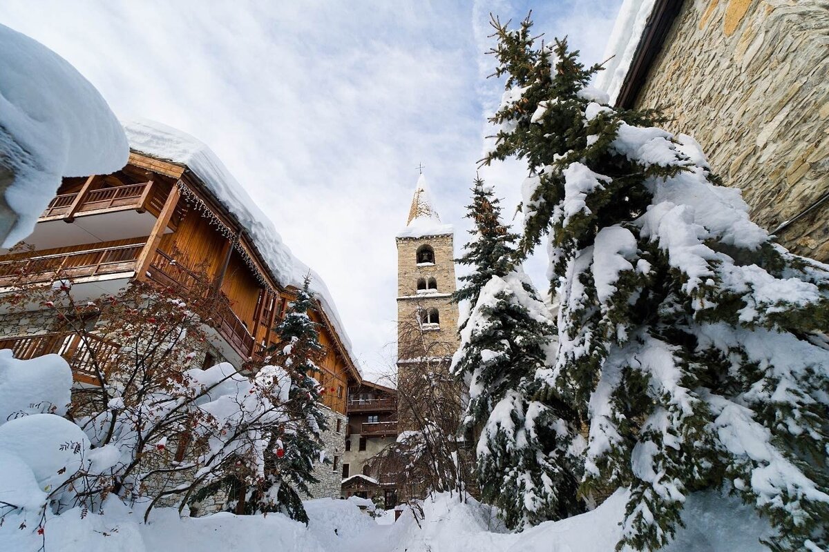 A quaint, snow-covered alpine village features a prominent stone church tower, wooden chalets, and frosted evergreen trees under a bright winter sky.