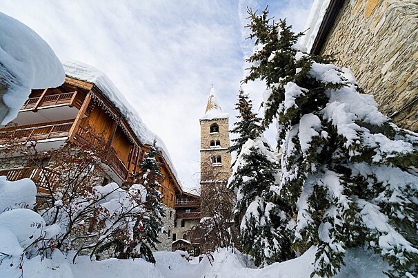 A quaint, snow-covered alpine village features a prominent stone church tower, wooden chalets, and frosted evergreen trees under a bright winter sky.