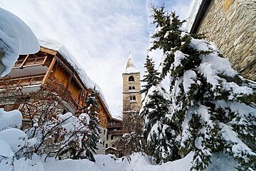 A quaint, snow-covered alpine village features a prominent stone church tower, wooden chalets, and frosted evergreen trees under a bright winter sky.