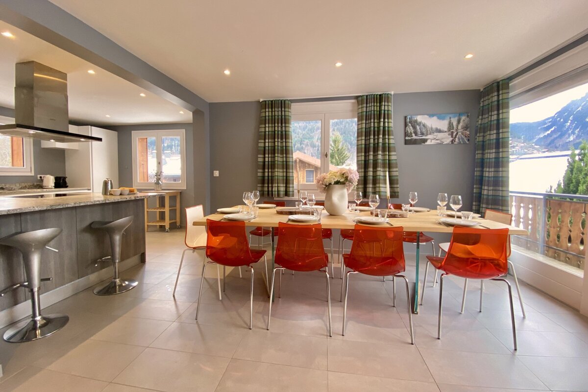 Spacious open-plan kitchen and dining area with a long wooden table, orange chairs, and modern bar stools, featuring a stunning mountain and lake view.