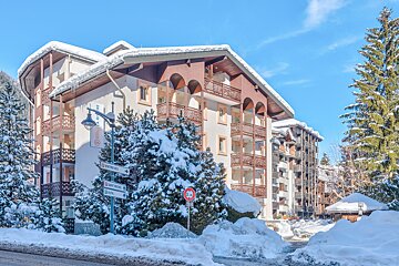 A snow-covered alpine building with balconies stands beside frosted pine trees and deep snow drifts under a clear blue sky.