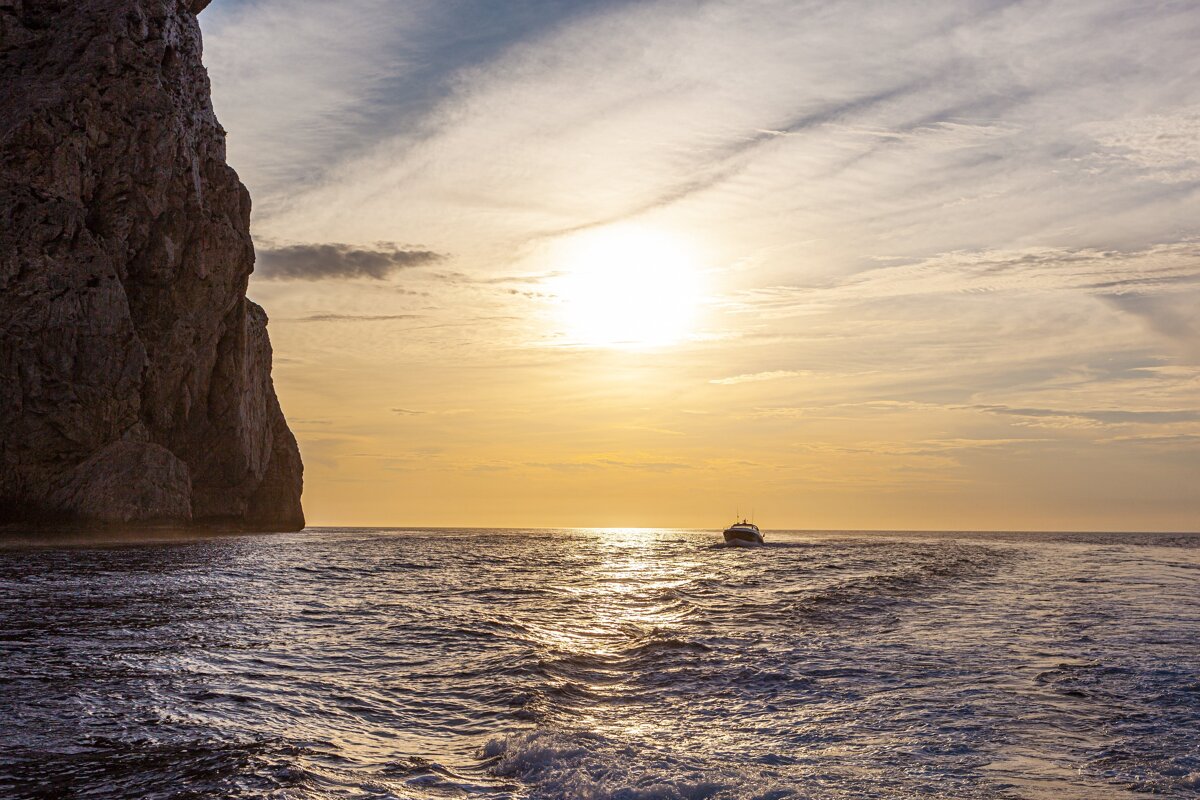 A boat sails away from a cliff into a golden sunset over wavy waters with dramatic skies.