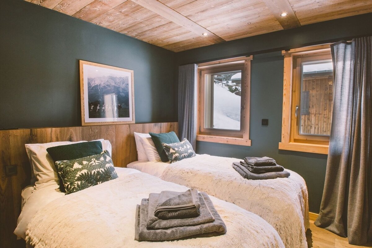 A cozy bedroom with two twin beds, dark green walls, wooden ceiling, and mountain views from windows. Soft bedding and towels complete the inviting space.