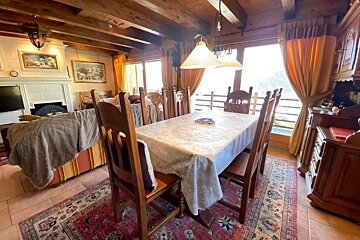 A cozy, rustic living-dining room with a wooden table, chairs, fireplace, beamed ceiling, and large windows offering a scenic view.