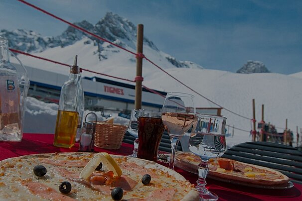 A sunny outdoor meal with pizza, wine, and water at a Tignes ski resort, featuring snow-capped mountains and a ski lift.