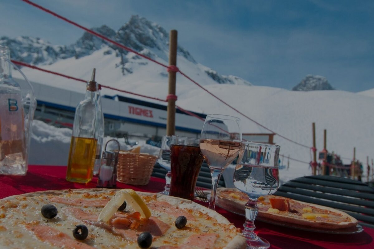 A sunny outdoor meal with pizza, wine, and water at a Tignes ski resort, featuring snow-capped mountains and a ski lift.