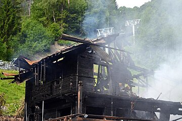 Smoke rises from a charred wooden building, likely a mountain lodge or restaurant, set against a green forest with ski lifts in the background.