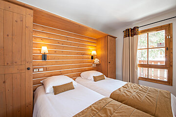 A cozy, wood-paneled bedroom with two single beds, warm lamps, and a window looking out to trees.
