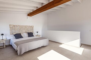 Bright bedroom with a carved headboard, white walls, exposed wooden beam, and sunlit tiled floor.