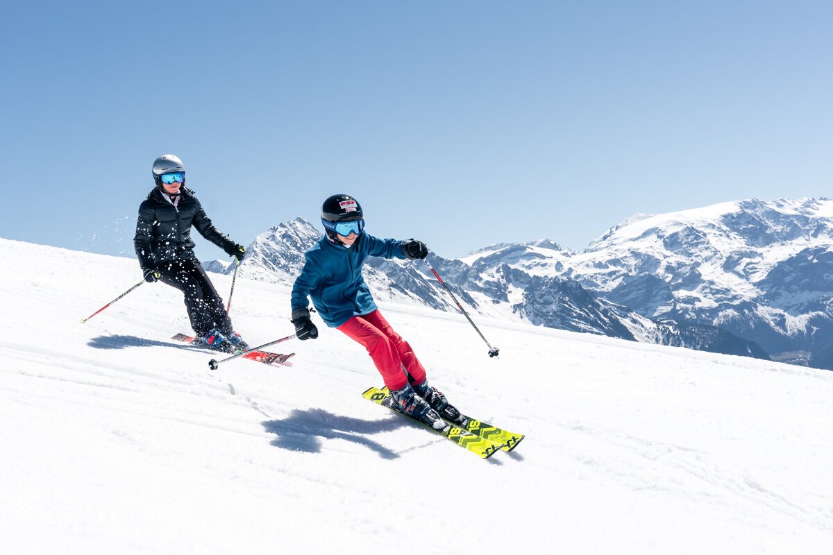 Two people ski down a bright, snowy mountain slope under a clear blue sky, with majestic snow-capped peaks in the background.