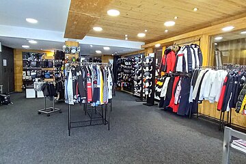Interior of a winter sports clothing store with racks of ski jackets, pants, and gear. Features wooden ceilings and a grey carpet.