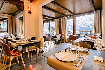 A cozy restaurant with modern decor, wooden beams, and set tables. Large windows overlook a snowy mountain landscape, creating a warm ambiance.