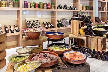A vibrant buffet table with terracotta bowls of anchovies, pickled vegetables, and eggs, alongside other dishes, against a backdrop of jarred goods on shelves.