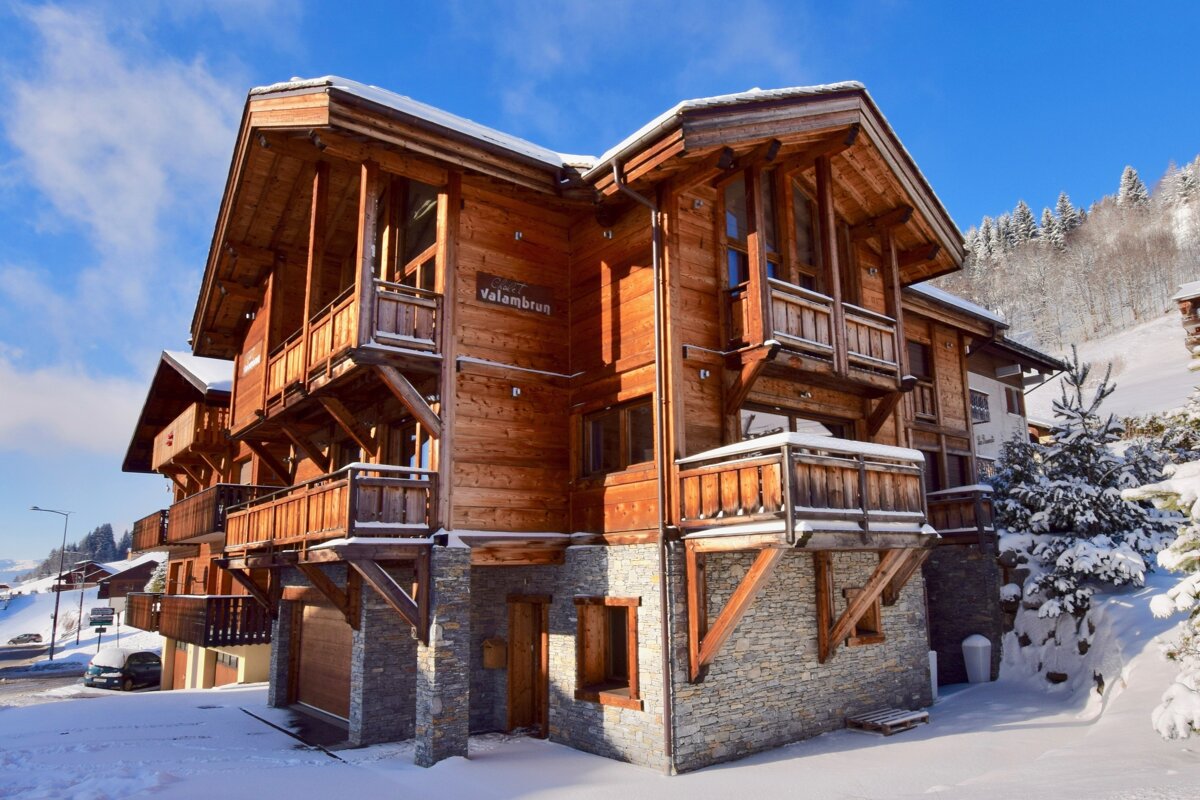 A beautiful multi-story wooden chalet with balconies and a stone base, covered in snow, under a bright blue sky in a winter mountain setting.