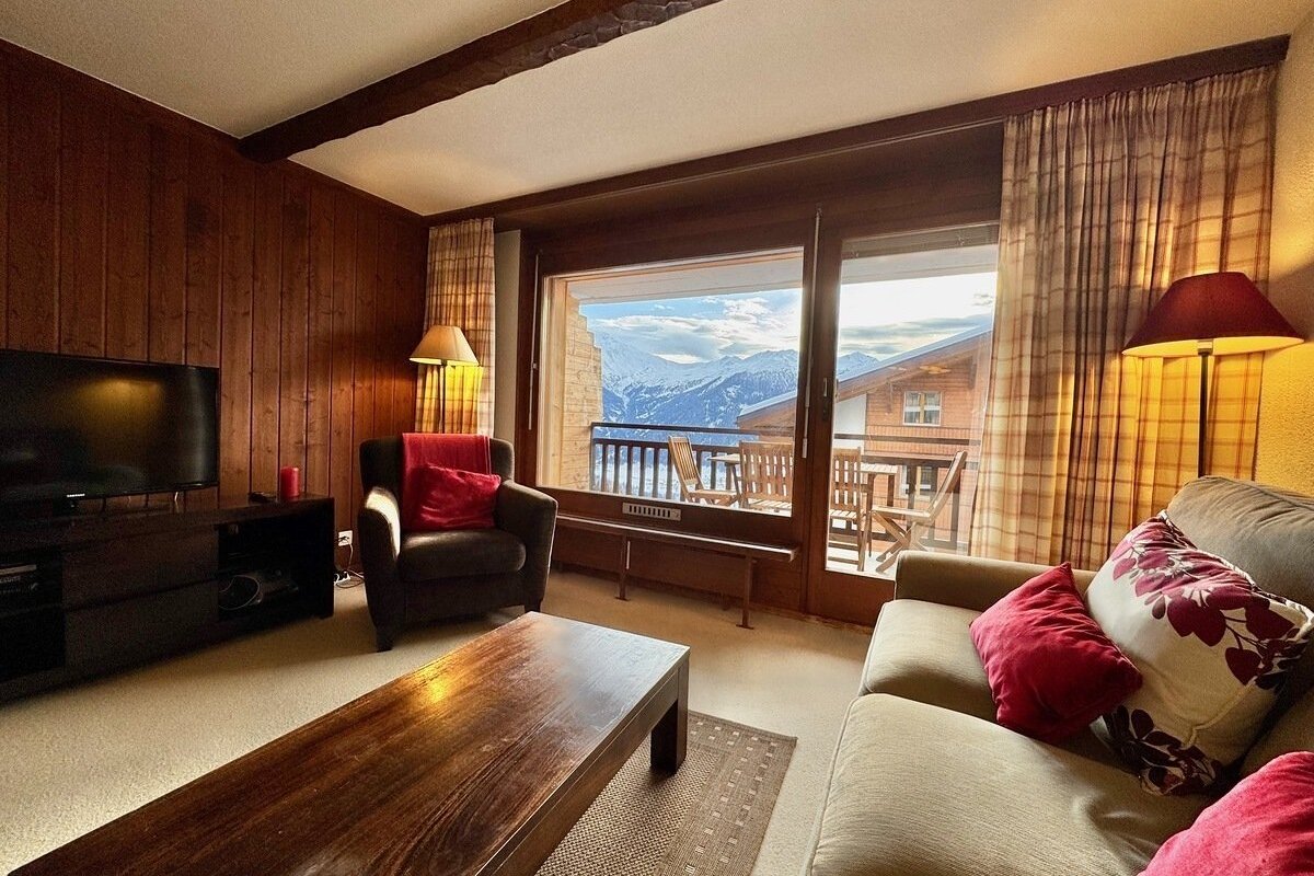 Wood-paneled living room with cozy furniture and a large balcony window revealing stunning snow-capped mountains.
