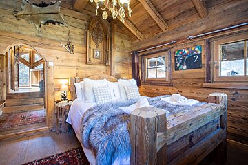 A cozy, rustic wooden bedroom features a large bed with fur throws, white pillows, and a chandelier. Windows frame the view, and an archway leads to a bright balcony.