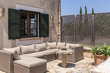 A sunny outdoor patio features a wicker sectional sofa, a stone table, and a potted plant next to an old building with green-shuttered windows.