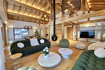 Spacious, modern chalet living room features high wooden ceilings, green sofas, a suspended fireplace, a Christmas tree, and a snowy outdoor view.