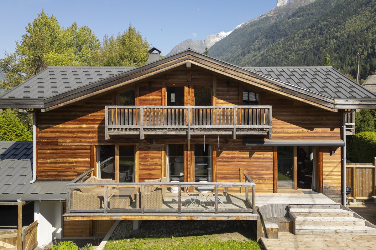 A rustic wooden chalet with balconies and decks sits against a backdrop of green trees and snow-capped mountains on a sunny day.