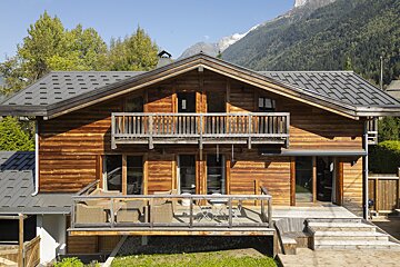 A rustic wooden chalet with balconies and decks sits against a backdrop of green trees and snow-capped mountains on a sunny day.