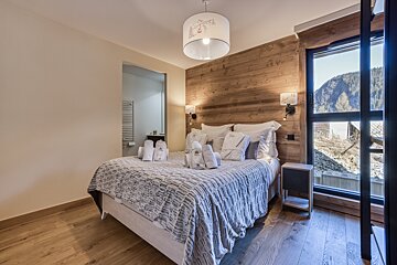 A cozy, modern bedroom featuring a large bed with white robes, wooden accent wall, and a tall window offering mountain views. A bathroom is visible.