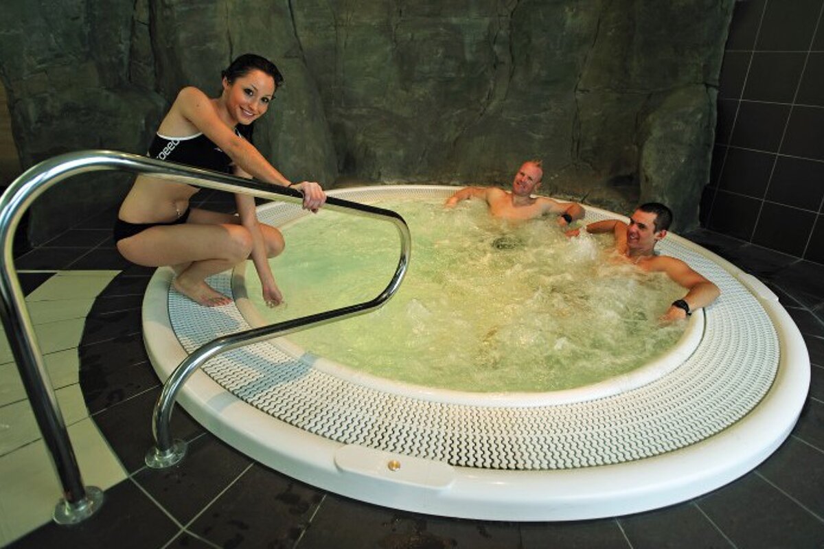 Two men and a woman are in a jacuzzi and the woman is wearing a swimsuit that says speedo