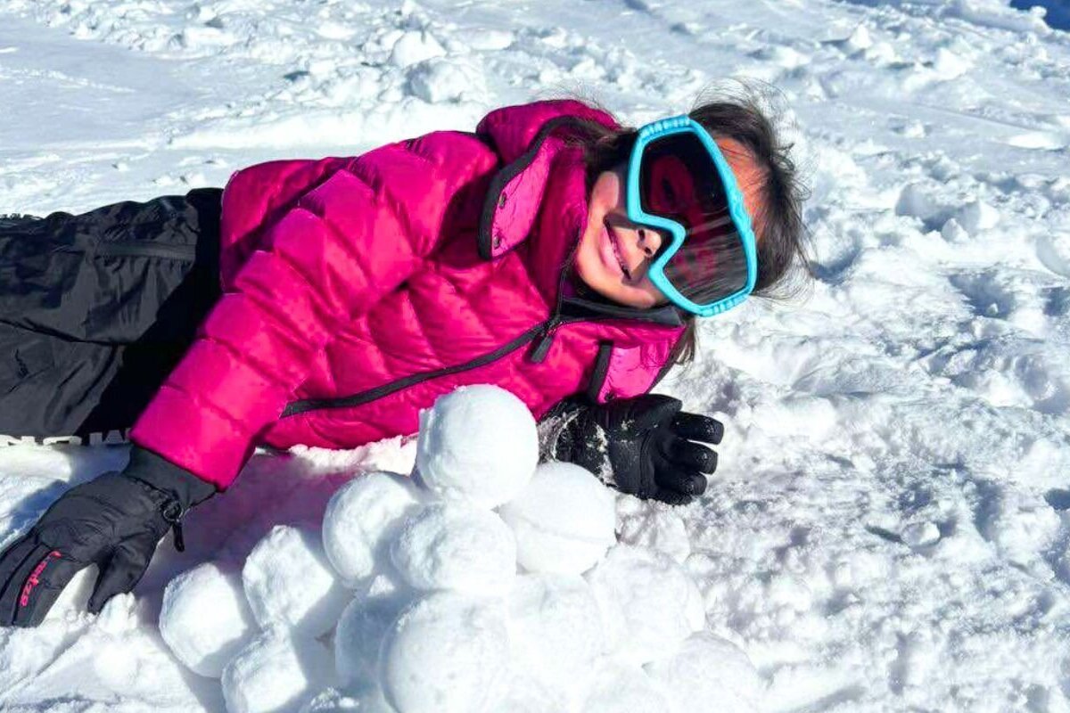 A smiling person in a pink jacket and blue goggles lies happily in sunny snow next to a pile of perfectly made snowballs.
