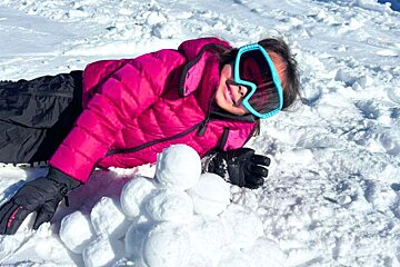 A smiling child in a pink jacket and blue goggles lies in the snow, next to a stack of snowballs.