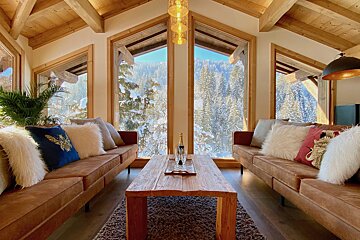 A warm chalet interior featuring two sofas, a wooden table with champagne, and large windows revealing a stunning snow-covered mountain forest.