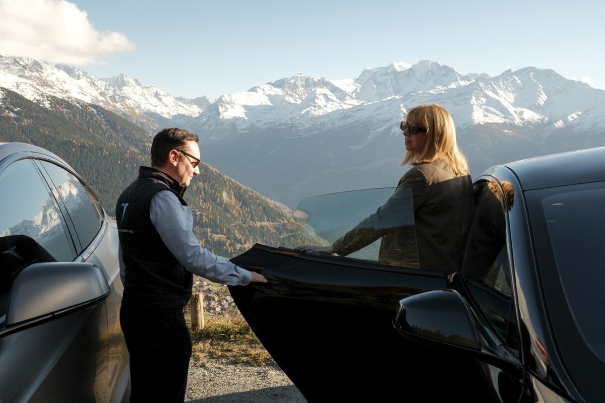 A man holds open a car door for a woman admiring a breathtaking view of snow-capped mountains and a valley from a high scenic point.