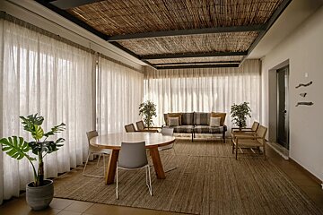 A bright, airy room featuring a woven bamboo ceiling, sheer white curtains, a natural rug, a round wooden table, and a striped sofa.