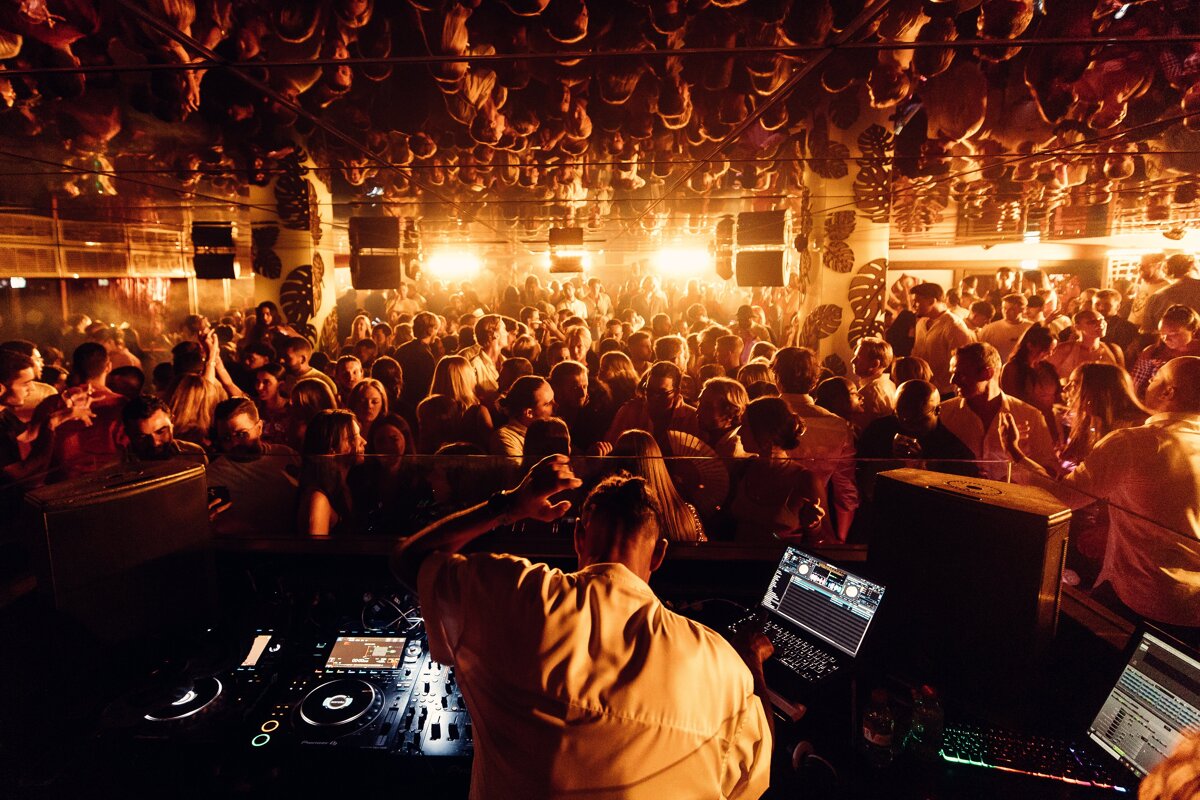 A DJ spins tracks for a packed, dancing crowd in a warm-lit nightclub, with reflections of the scene visible on the ceiling.