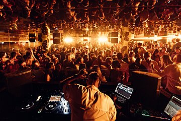 A DJ spins tracks for a packed, dancing crowd in a warm-lit nightclub, with reflections of the scene visible on the ceiling.