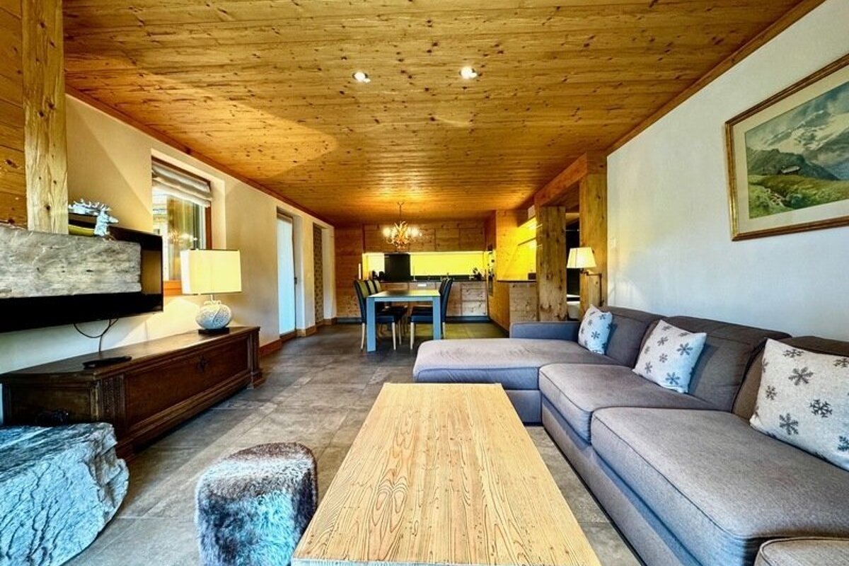A cozy, rustic alpine living room featuring wood ceilings, a large grey sectional, wooden coffee table, and an adjacent dining area.