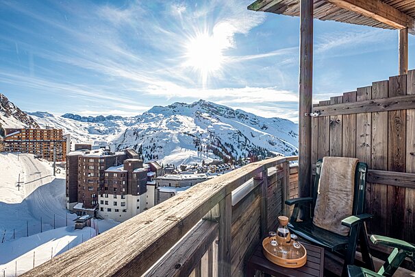 A sunny wooden balcony overlooks a stunning snow-covered mountain range and ski resort village under a bright blue sky.