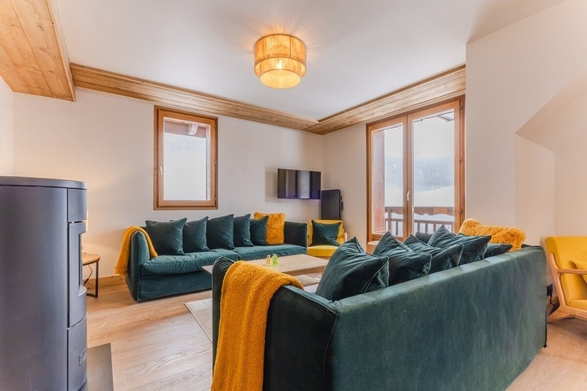 Cozy cabin living room featuring dark green velvet sofas with mustard yellow throws, wooden accents, and large windows.