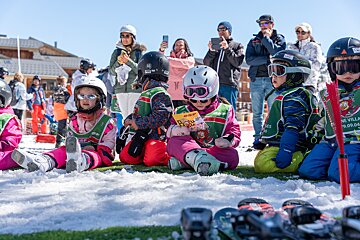 Ski school kids in helmets & goggles sit on snowy grass, watched by adults. One reads a pamphlet, with skis in the foreground.